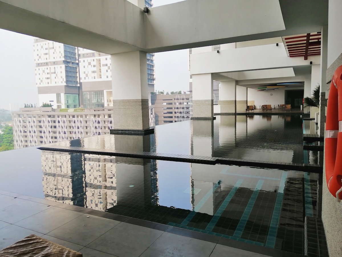 An infinity pool is showcased, reflecting the surrounding architecture and greenery. Smooth tile flooring leads to the water's edge, with seating areas visible in the background. The space is brightened by ample natural light entering through the large openings above.