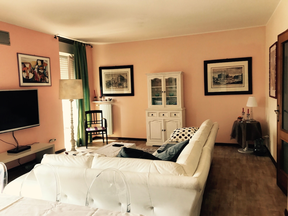 A spacious living room is shown, featuring a large white sofa and a coffee table. A flat-screen TV is mounted on the wall, while framed artwork adorns the walls. A tall lamp and a small side table with decorative items add warmth to the space.