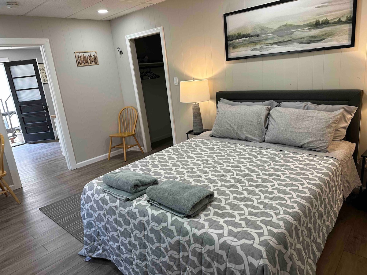 A comfortable sleeping area is highlighted by a queen-size bed, adorned with gray patterned bedding and neatly arranged towels. Two nightstands, each with a lamp, flank the bed, while a wooden chair offers additional seating. A closet provides storage space, and art decorates the wall.