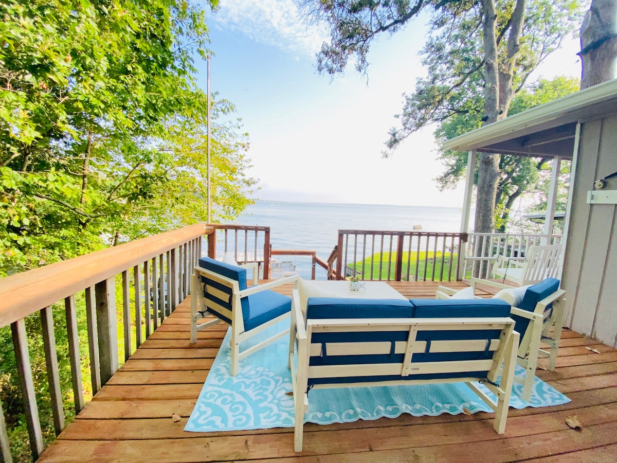 An outdoor deck features a set of light blue cushioned chairs arranged around a small table. The deck overlooks a tranquil lake, with surrounding greenery and trees enhancing the peaceful setting. A soft blue area rug is placed beneath the seating arrangement.