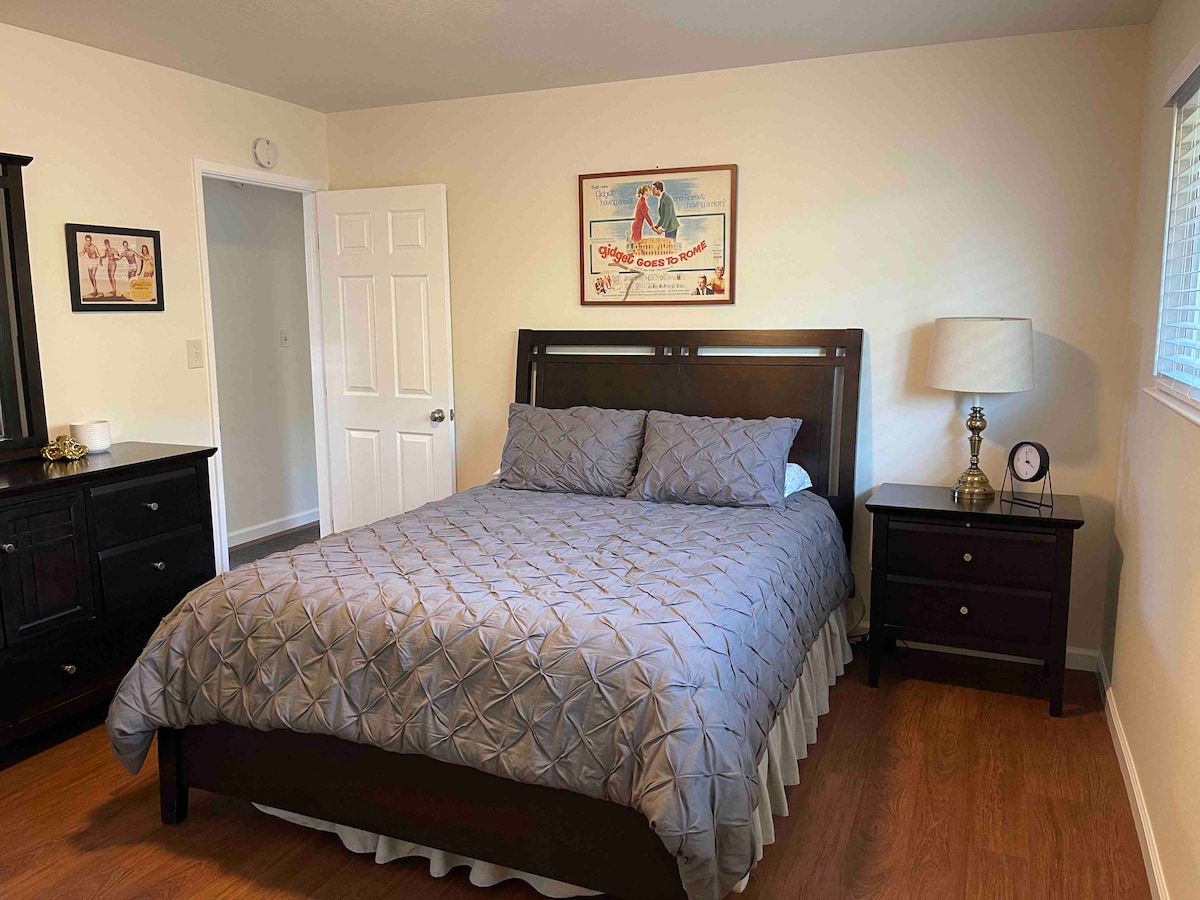 A tranquil bedroom is featured, showcasing a spacious bed with a geometric quilt and matching pillows. A wooden bedside table holds a lamp and a clock. Complementing furniture includes a dresser, and a framed vintage art piece decorates the wall above the bed.