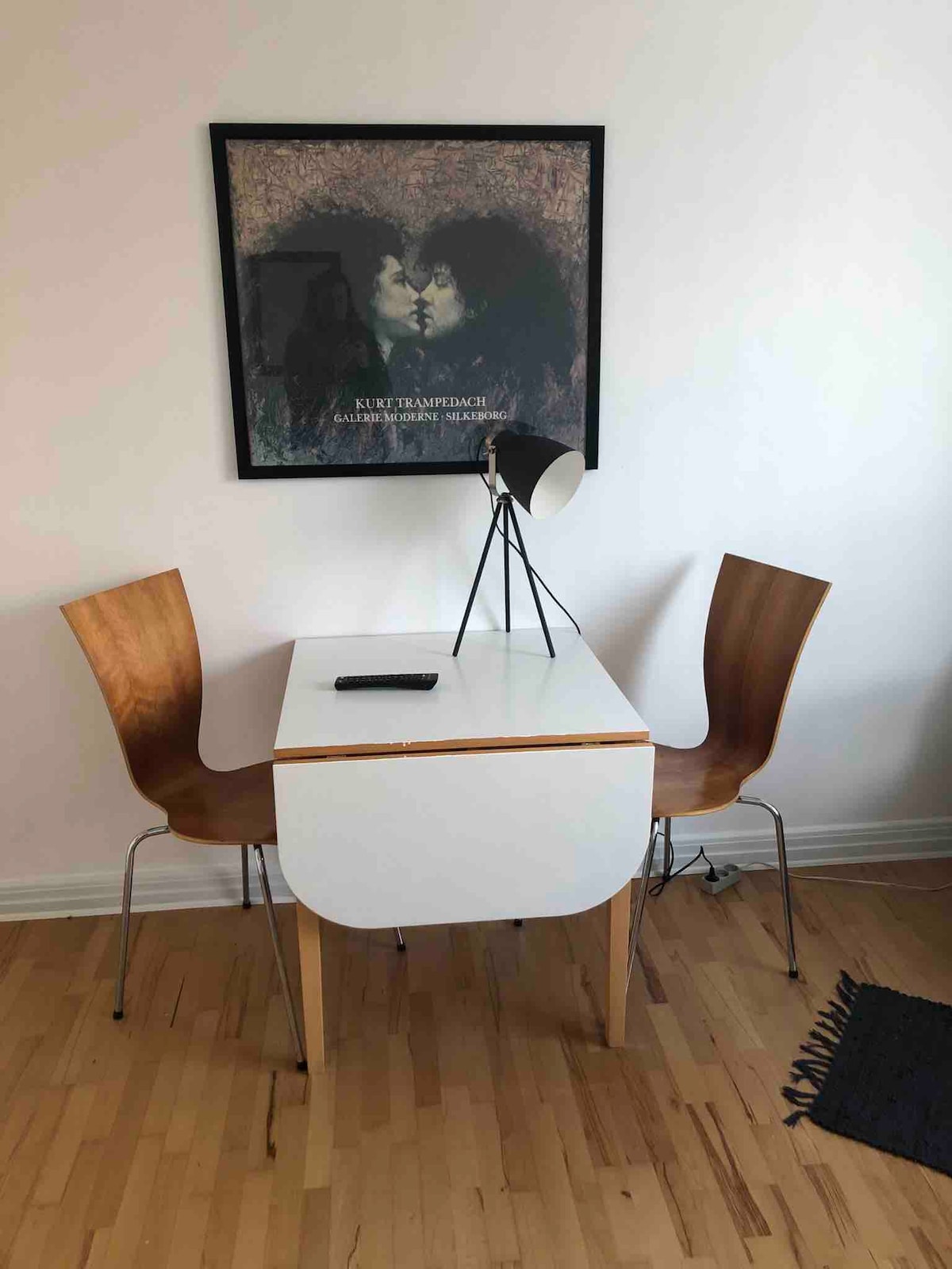 A simple dining table with a rounded edge is accompanied by two wooden chairs. A sleek lamp stands on the table, while a framed artwork is mounted on the wall above. The smooth wooden floor adds to the minimalistic design.