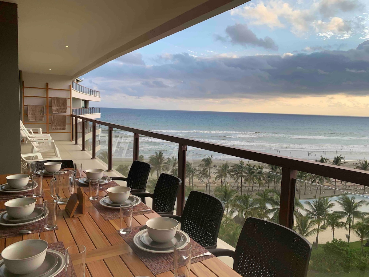 A spacious balcony features a wooden dining table set for six, accompanied by elegant dinnerware. The view overlooks the beach and ocean, with waves gently lapping at the shore. Lush palm trees line the beach, creating a serene coastal backdrop under a cloudy sky.