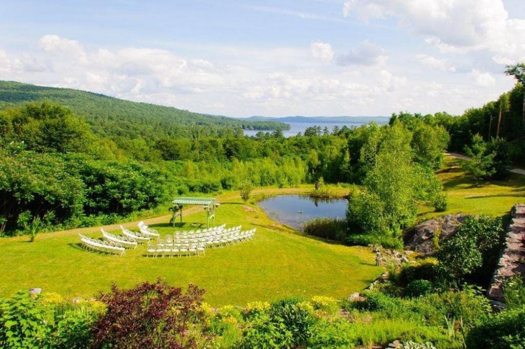An expansive outdoor space is set up for events, featuring white chairs arranged in rows facing a simple arch. A pond is nestled in the lush green landscape, bordered by trees and wildflowers. Newfound Lake and distant mountains provide a scenic backdrop.