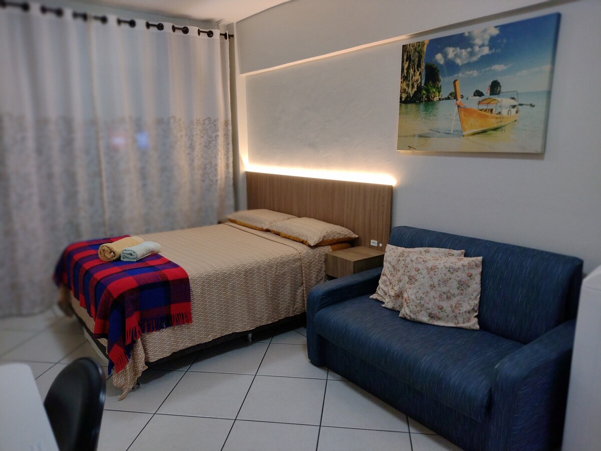A loft-style studio is shown with a spacious bed dressed in neutral linens, accompanied by a colorful blanket. A blue sofa with floral cushions is visible beside the bed. Soft lighting highlights the decor, and sheer curtains allow natural light to filter in.