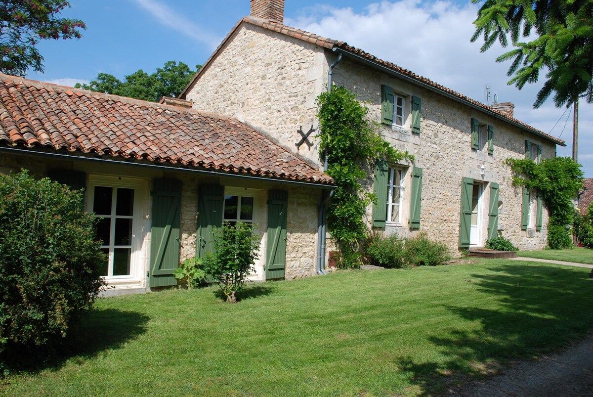A charming stone farmhouse is set amidst lush green gardens. The structure features a classic tiled roof, wooden shutters in shades of green, and large windows allowing natural light. The well-maintained lawn provides a spacious area for relaxation.