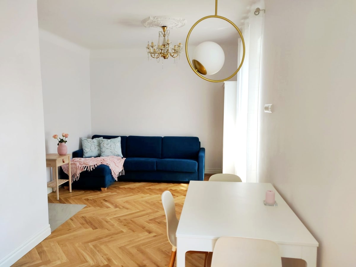 A well-lit living area displays a blue sofa adorned with light decorative pillows. A round mirror is suspended from the ceiling, reflecting natural light. A small dining table with chairs occupies one corner, enhancing the space's functionality. Light wood flooring adds warmth to the room.