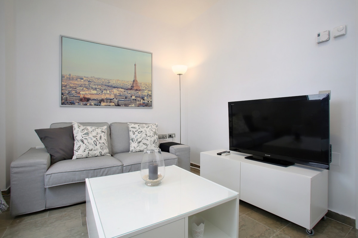 The living area features a comfortable gray sofa with decorative pillows, a sleek white coffee table, and a flat-screen TV. A large wall-mounted artwork depicts a cityscape, while an elegant floor lamp provides soft illumination, creating a welcoming space for relaxation.