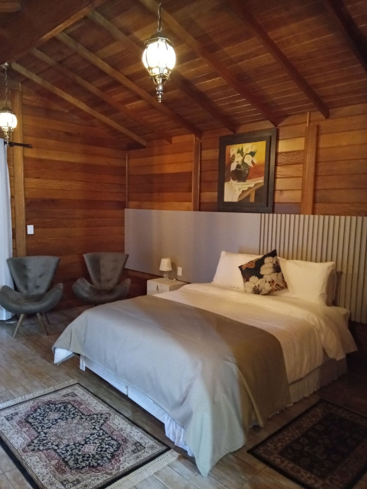 A cozy bedroom features warm wooden paneling and beams. A spacious bed is dressed in neutral linens, accompanied by decorative pillows. Two armchairs are positioned near a bedside lamp, and a piece of artwork adorns the wall, adding a subtle touch of style.