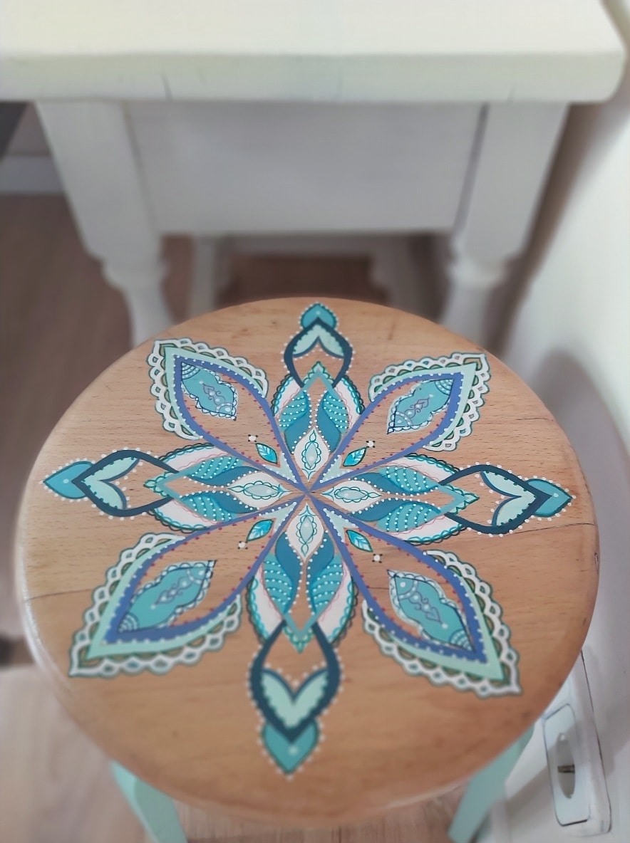 A wooden stool features a detailed, hand-painted mandala design in shades of blue and green. The intricate patterns radiate from the center, showcasing an artistic flair. The stool is positioned next to a light-colored table in a well-lit space.