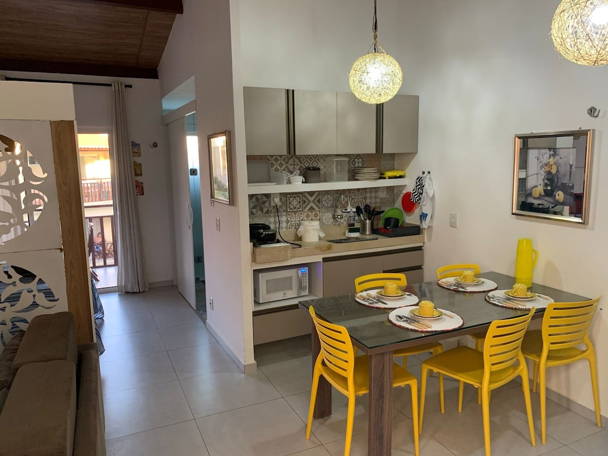 A modern kitchen area features sleek cabinetry and stainless-steel appliances, including a microwave. A wooden dining table with yellow chairs is set for four, showcasing colorful dishware. Natural light is provided by a nearby window, creating a cheerful atmosphere.