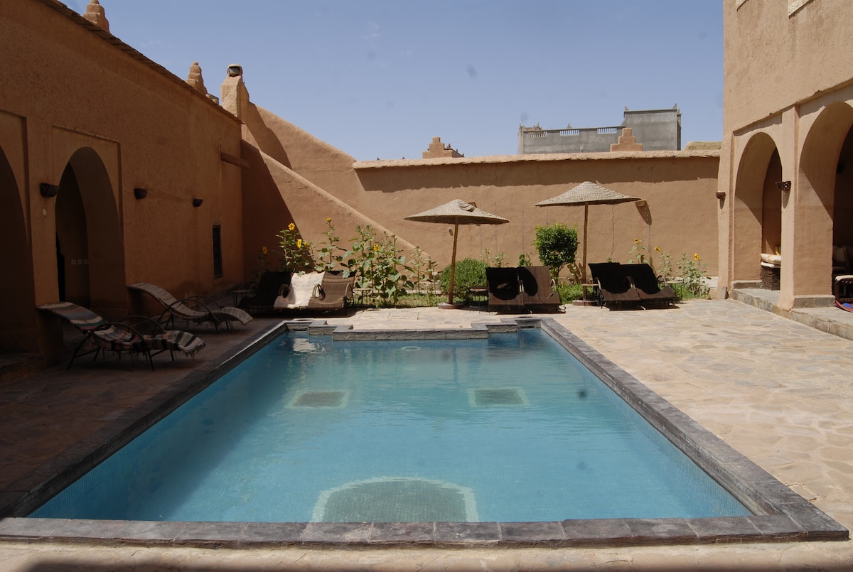 A tranquil courtyard features a clear pool surrounded by stone paving. Two umbrellas provide shade over comfortable loungers, while lush greenery enhances the serene environment. Arched doorways can be seen in the background, connecting to the surrounding structures.
