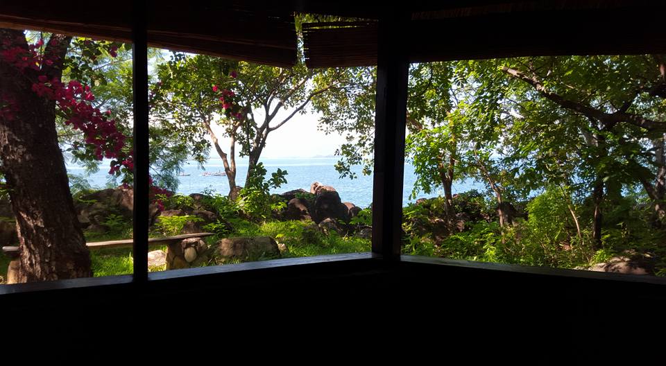 The view from a large open window frames vibrant greenery, featuring flowering plants and rocky terrain leading to the lake. The calm waters of the lake reflect the surrounding nature, providing a serene backdrop to the picturesque setting.