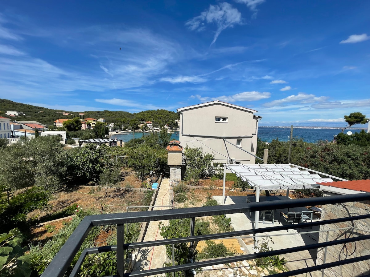 A sweeping view from a balcony reveals a lush garden and a nearby beach, framed by distant hills. The landscape is punctuated by houses, olive trees, and a pergola offering shaded outdoor seating. The vibrant sky contributes to a serene atmosphere.