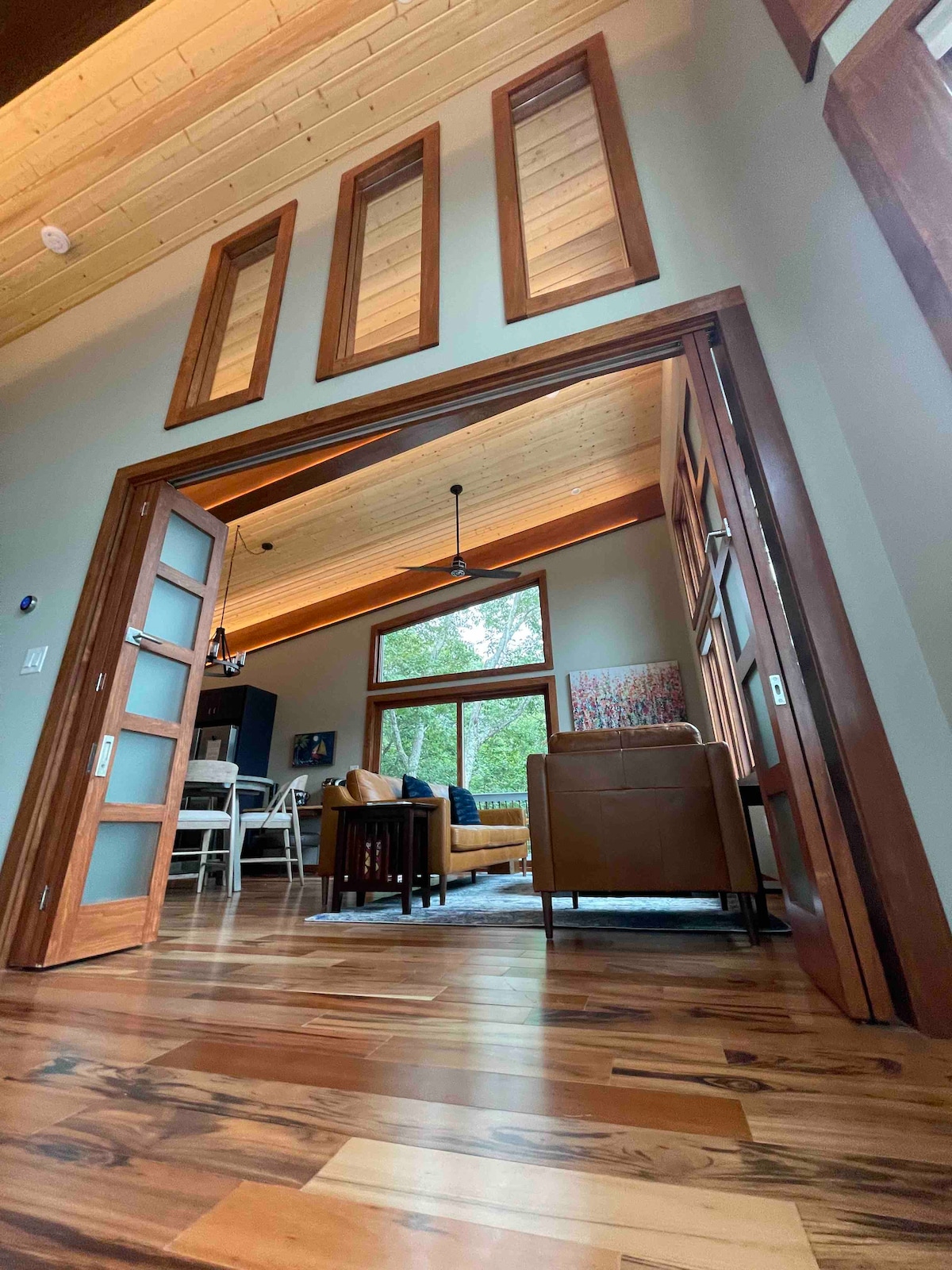 A view from the entrance capturing the spacious living area. Natural light floods through large windows, highlighting the warm-toned tigerwood flooring and wood ceiling. The open layout connects the living space with dining and kitchen areas, showcasing a modern and inviting design.