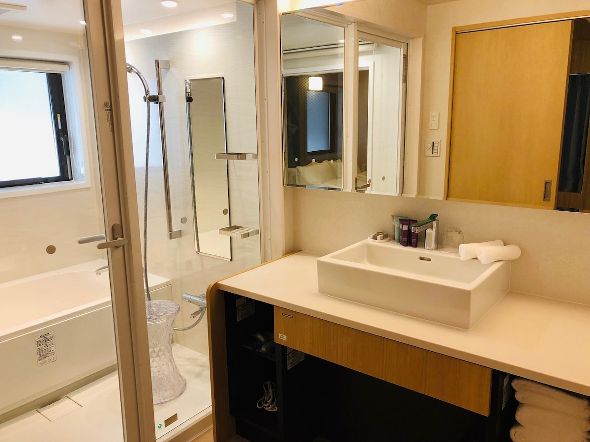 A spacious bathroom features a glass-enclosed shower and a separate bathtub. A large vanity with a sink is complemented by a mirror and wooden cabinetry. Natural light enters through a window, enhancing the functional layout that includes neatly arranged towels and toiletries.