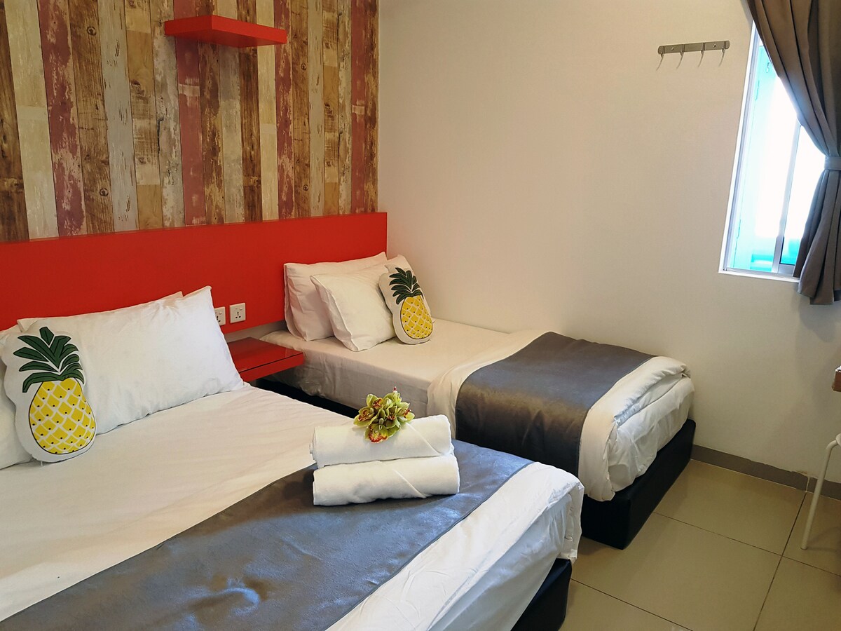 The room contains two beds with soft linens, accented by decorative pineapple cushions. A vibrant red headboard complements the rustic wall, while natural light enters through a window with a curtain. A small shelf is positioned above the headboard, adding functional decor.