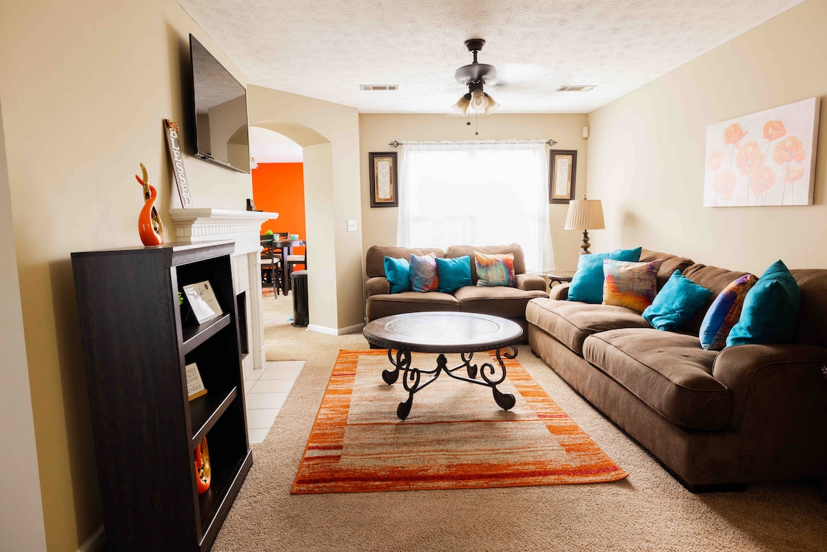 A welcoming living area features a spacious sectional sofa adorned with colorful cushions. A round coffee table sits centrally on a patterned area rug, while soft light filters in through a window draped with sheer curtains. A television is mounted on the wall.