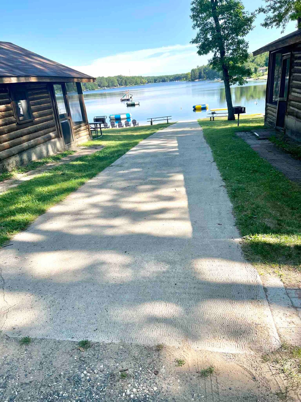 A concrete pathway lined with greenery leads towards the lake, flanked by rustic cabins on either side. The lake is visible in the background, featuring colorful chairs and a floating water trampoline. Soft sunlight filters through the trees, creating a serene atmosphere.