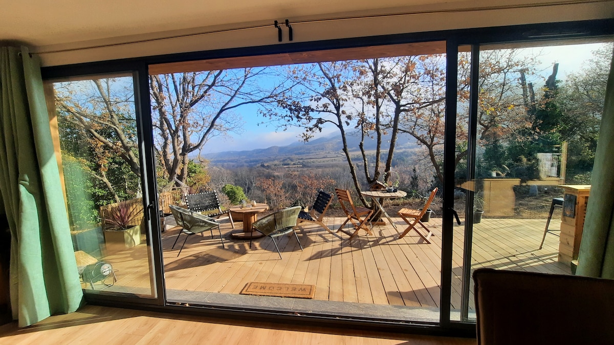 A large glass sliding door offers a view of a spacious outdoor terrace with a mix of seating options, including chairs and a table. The surrounding trees provide a natural backdrop, while distant mountains and valleys extend in the horizon, reflecting a tranquil outdoor environment.