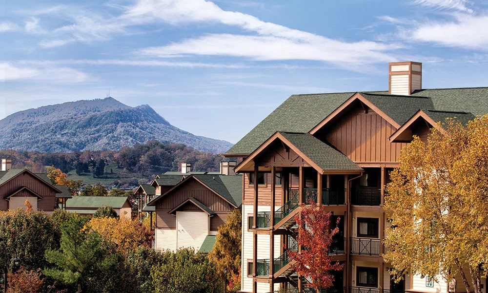 The image showcases a multi-story resort building nestled among vibrant autumn foliage. Mountains are visible in the background under a clear blue sky, while the architecture features a combination of wood and stone elements, with a peaked roof and balconies.