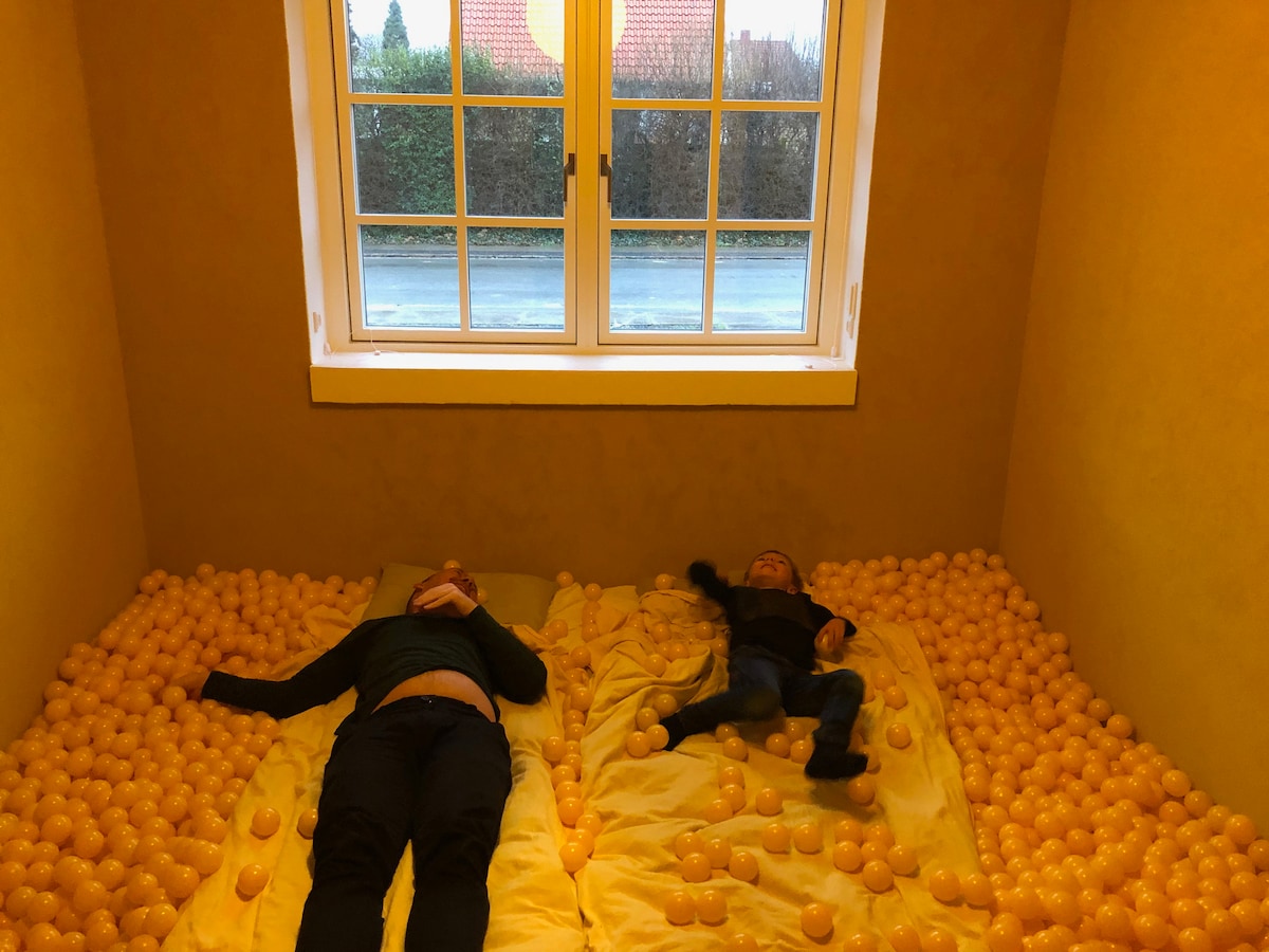 A playful room is filled with vibrant yellow plastic balls, covering the floor and part of the bed. Two people are lying down, relaxed, amidst the colorful balls. A window is visible, allowing natural light to fill the space and showcase the cheerful atmosphere.