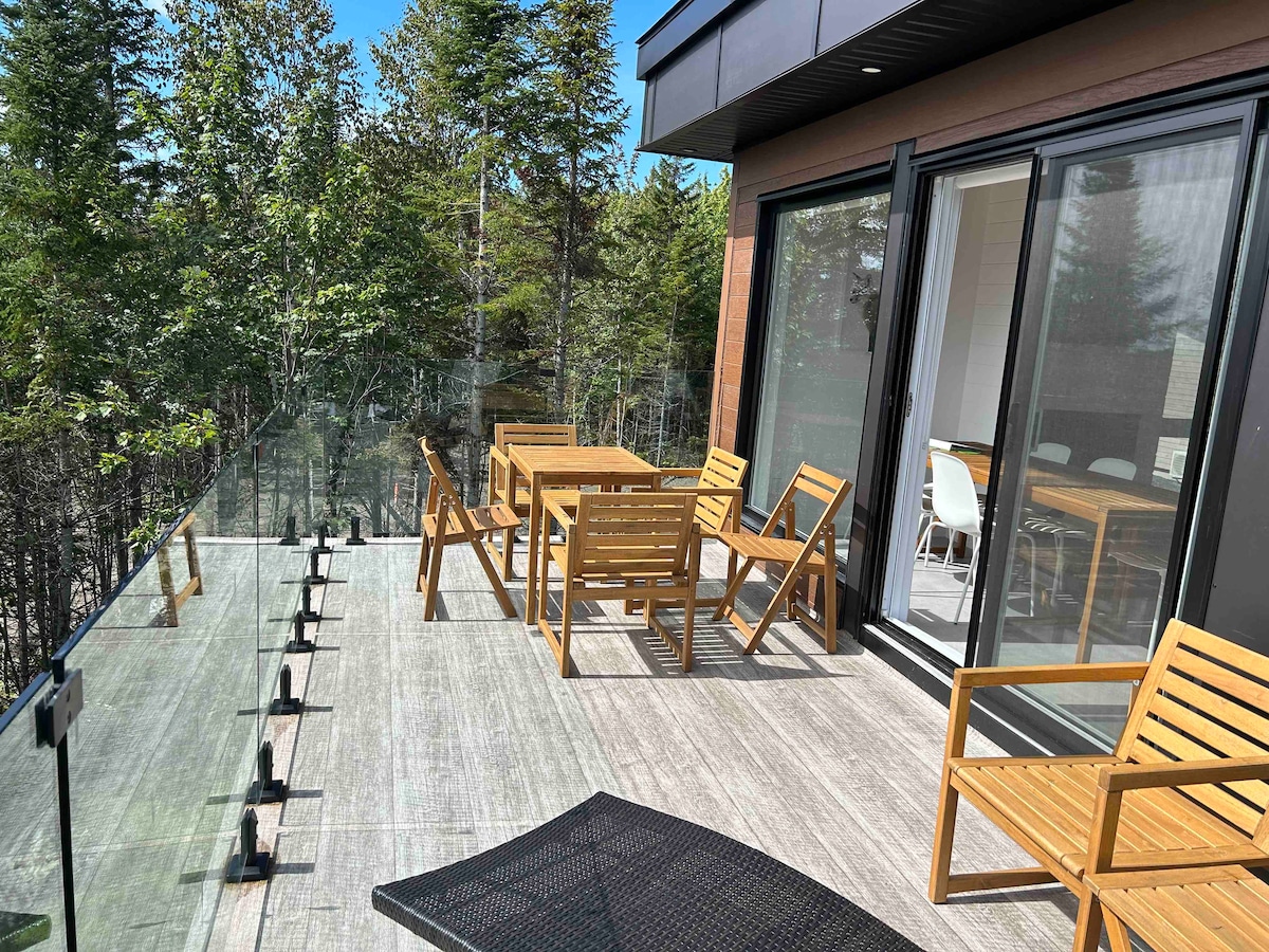 An outdoor terrace features wooden furniture, including a dining table and seating chairs. Glass railings provide unobstructed views of the surrounding trees and nature, while sunlight illuminates the deck, creating a bright and inviting space for relaxation.