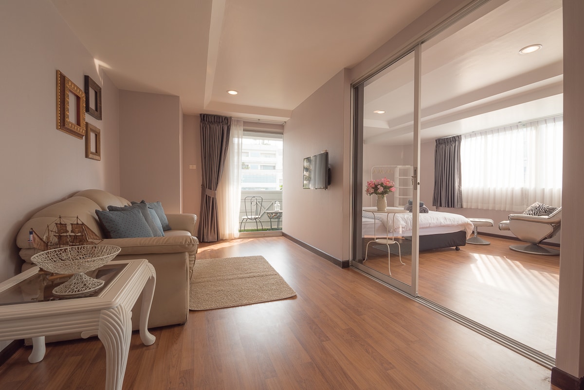 The image depicts a modern apartment interior featuring a cozy sitting area with a light-colored leather sofa. Natural light floods the open space, leading to a bedroom beyond, where a spacious bed and window with sheer curtains are visible. A small balcony can also be seen.
