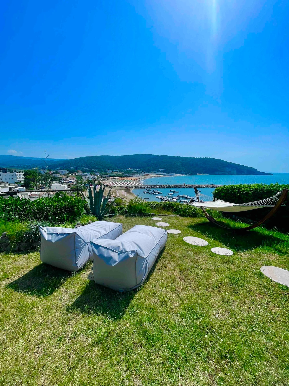 A well-maintained garden area features two light-colored lounge chairs positioned on green grass. A hammock is visible nearby, offering a relaxing space with views of the blue waters and beach beyond. The vibrant landscape is complemented by distant hills and a clear blue sky.