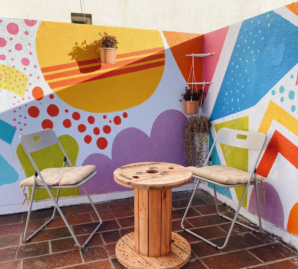 A vibrant outdoor patio space is presented, featuring a colorful mural with geometric shapes. Two folding chairs with cushions are positioned beside a wooden spool serving as a table. Small plants can be observed in pots attached to the wall.