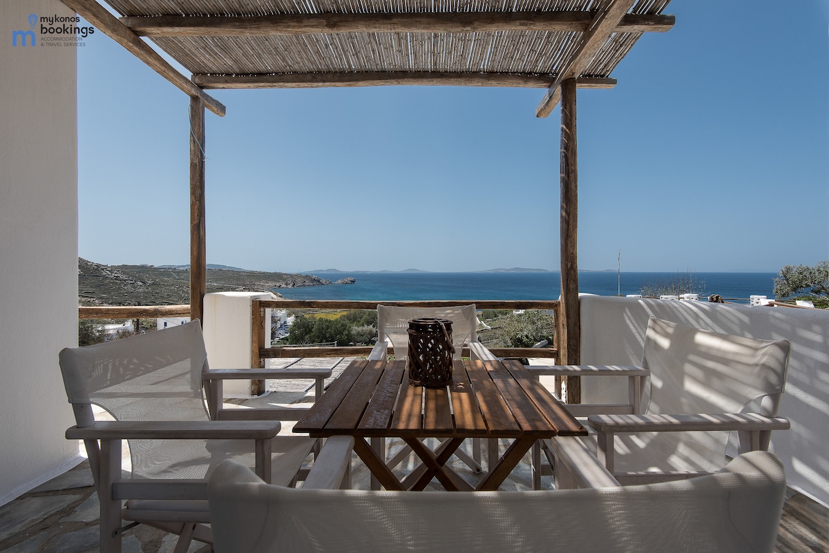 A spacious balcony area features a wooden table surrounded by four white chairs, ideal for relaxation. The sea view is visible in the background, framed by a wooden pergola that provides shade, enhancing the outdoor seating experience.