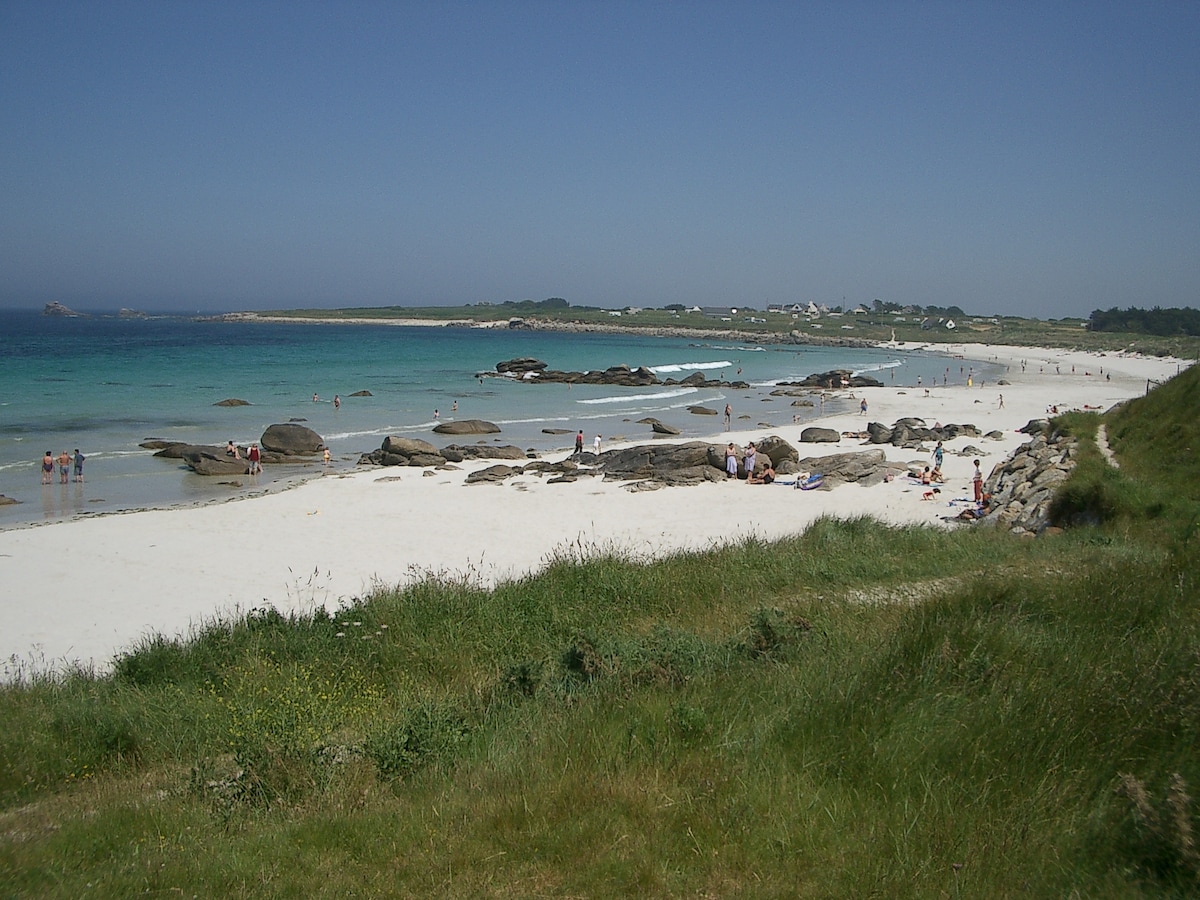A sandy beach extends into the distance, framed by gently rolling green hills. Sunbathers and families are scattered along the shoreline, with shallow turquoise waters lapping at the pale sand. Rocky formations are visible near the water's edge, adding texture to the landscape.