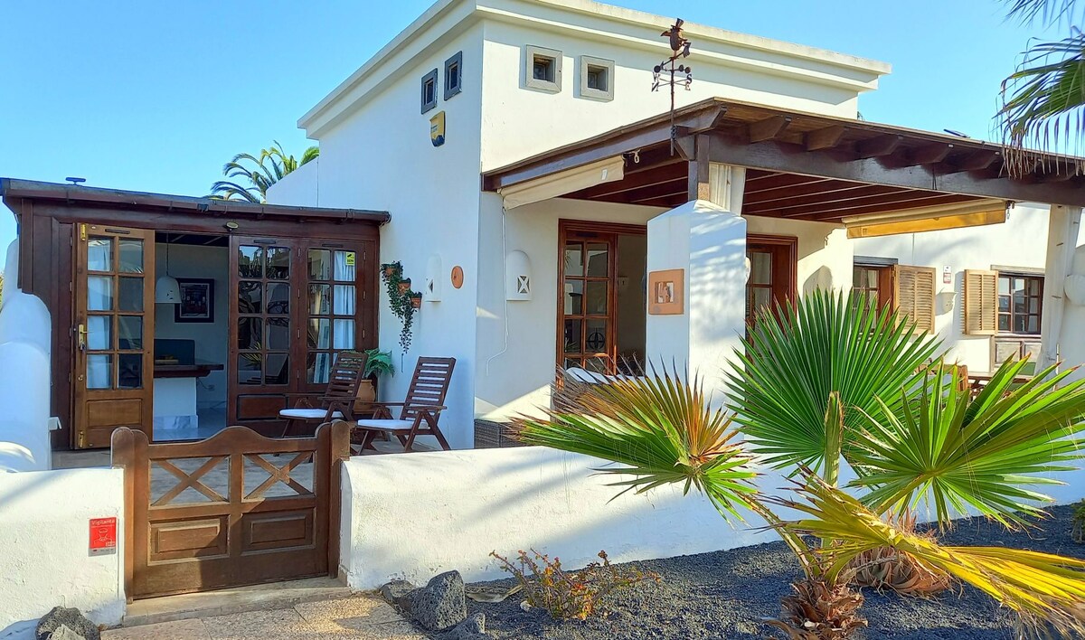 The exterior of the villa showcases a charming white structure with wooden accents. A gated entrance leads to a landscaped area featuring palm trees and volcanic soil. Relaxing lounge furniture is positioned on a covered patio, inviting guests to enjoy the outdoor space.