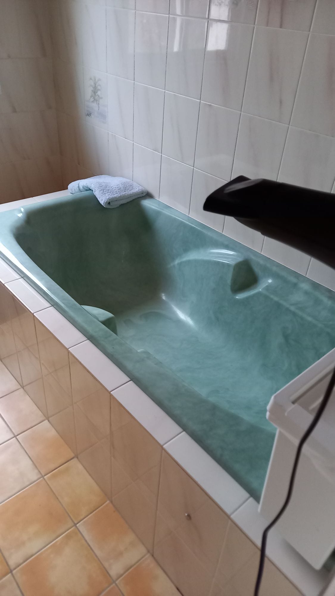 A spacious bathtub is featured, showcasing a green marble design. A folded towel is placed on the edge, and a hairdryer is positioned nearby. The tiled walls and floor reflect a neutral color palette, enhancing the bathroom setting.