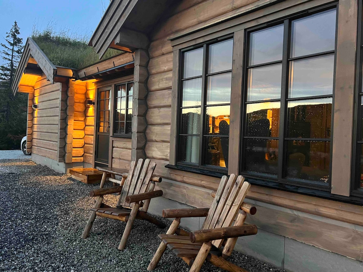A rustic log cabin exterior features large wooden windows, allowing natural light to fill the interior. Two comfortable outdoor chairs are positioned on a gravel area, inviting relaxation. The warm glow from the cabin's exterior lights adds a welcoming touch during twilight.