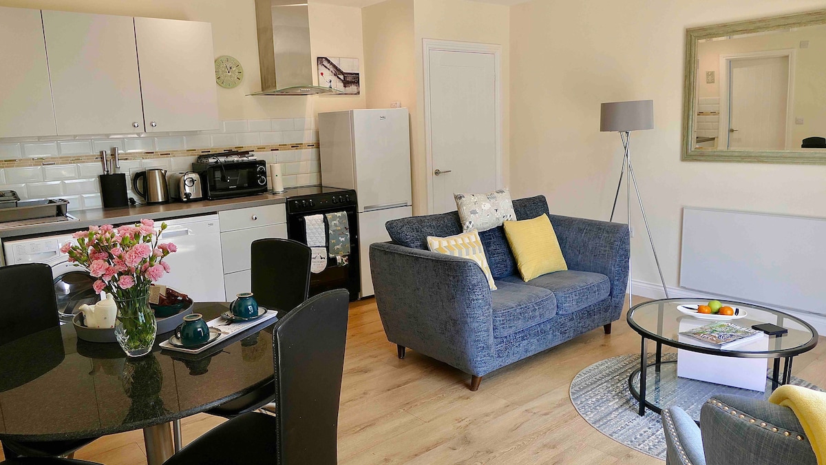 A well-equipped living space includes a cozy blue sofa paired with patterned cushions. A circular coffee table sits on a light rug, surrounded by a dining table set for two. The kitchenette features modern appliances, while natural light fills the room from the windows.