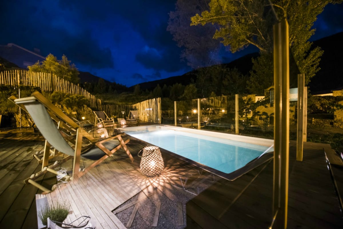 An outdoor swimming pool is surrounded by wooden decking and elegant lounge chairs. Soft lighting from lanterns creates a calming atmosphere, while the dark evening sky and distant mountains enhance the tranquil setting.