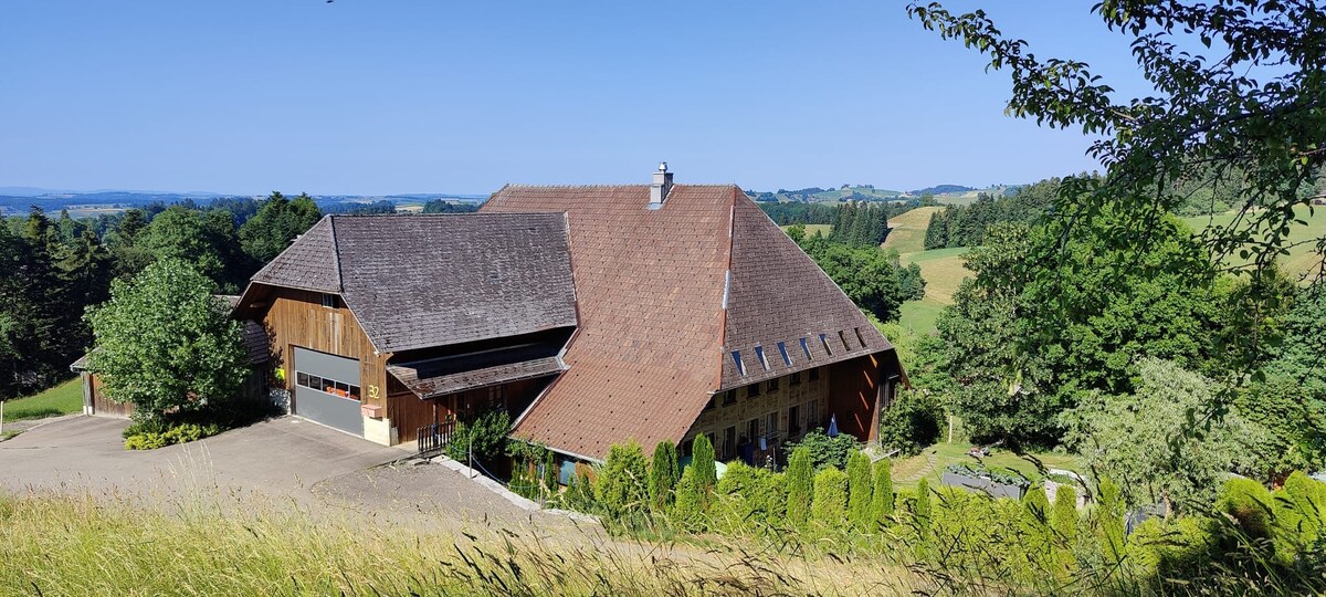 A spacious countryside building with a large sloped roof sits on a gentle hillside, surrounded by lush greenery and open fields. Large garage doors are visible on one side, and a well-maintained garden area is apparent at the front, enhancing the serene landscape.