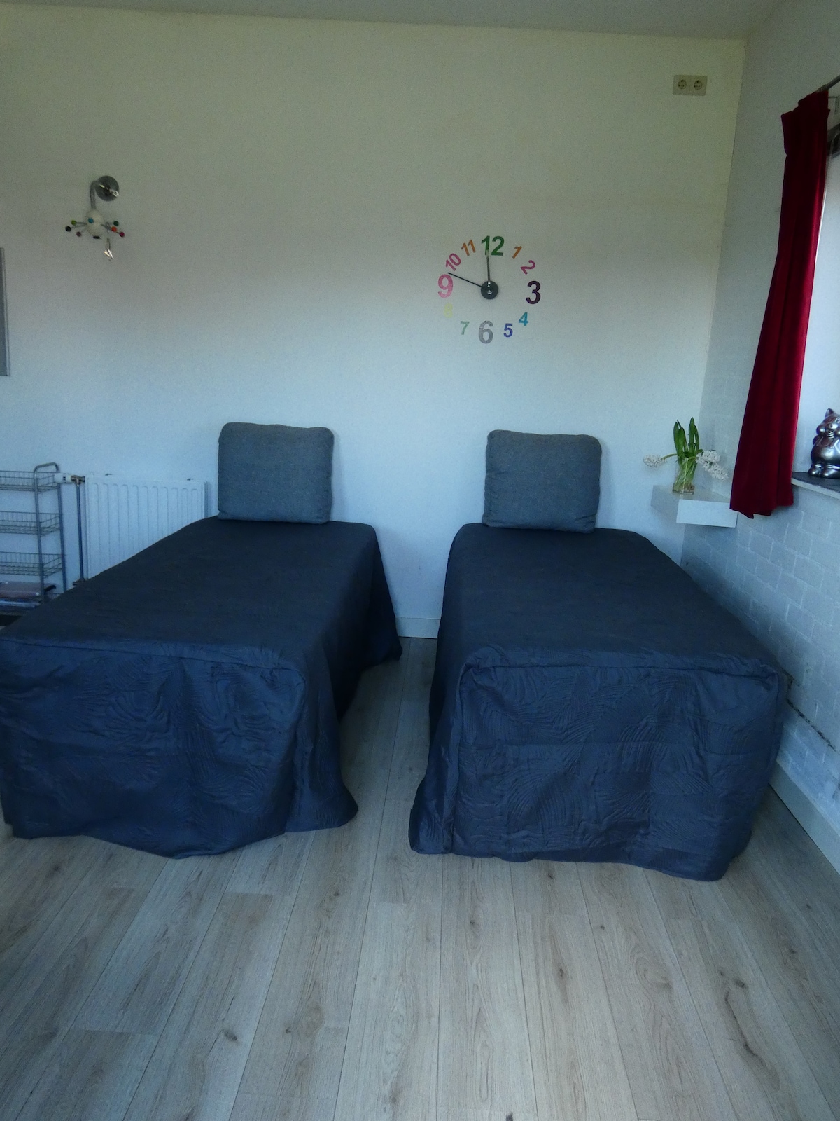 The image features a tranquil sleeping area with two single beds covered in dark grey bedspreads. Each bed is paired with a simple headboard and a grey cushion. A wall clock is displayed above, showing the time clearly, while a window with a red curtain allows natural light to enter.