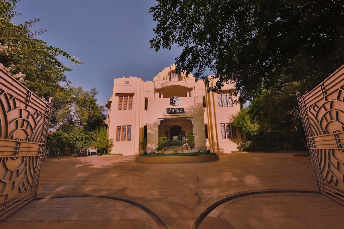 The heritage Chettiar mansion presents a grand façade with art-deco elements, framed by lush greenery. The entrance is marked by ornate gate designs and a circular driveway leading to the main entry, inviting exploration of its historical architecture.
