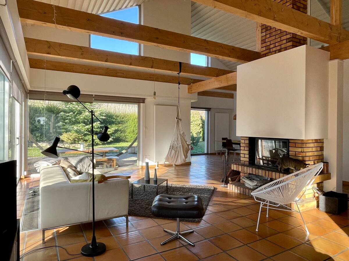 The spacious living area features wooden beams and large windows that allow natural light to flood in. A cozy seating arrangement is arranged around a fireplace, complemented by a standing lamp. A hammock hangs near the windows, offering a relaxed connection to the garden.