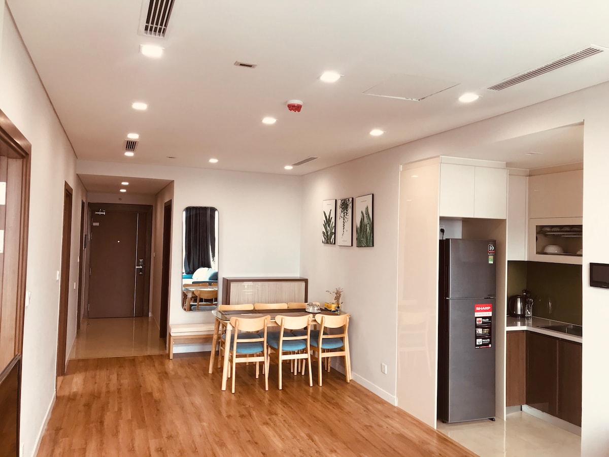 An open living space features a dining area with a wooden table and six chairs, positioned alongside a sleek kitchenette. The walls are decorated with botanical prints, and natural light fills the room, enhancing the wooden flooring and modern furnishings.