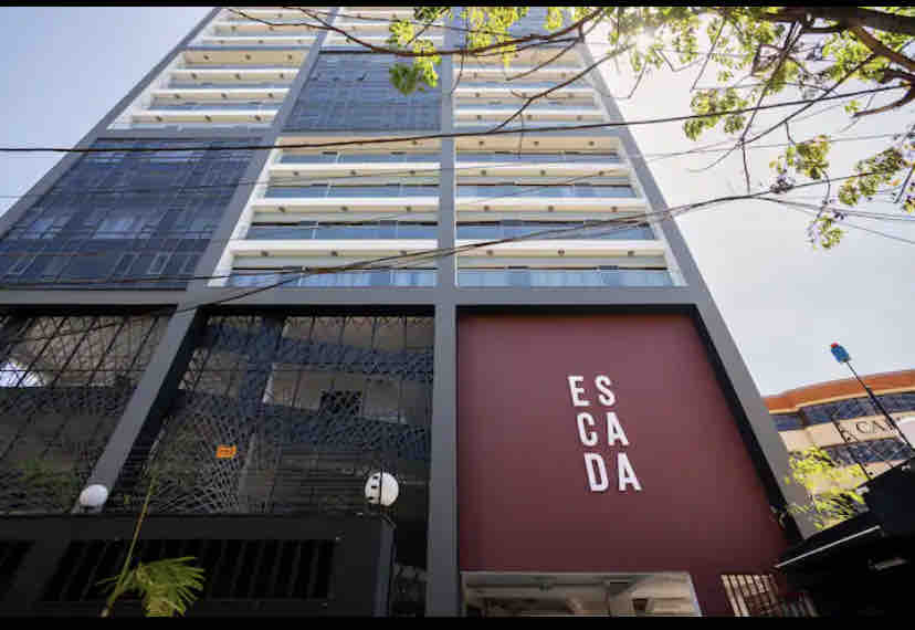 A modern multi-story building's exterior is depicted, featuring a contemporary design with large glass panels. The prominent building name, 'ESCADA,' is displayed on a bold maroon wall at the entrance, complemented by green foliage near the base.
