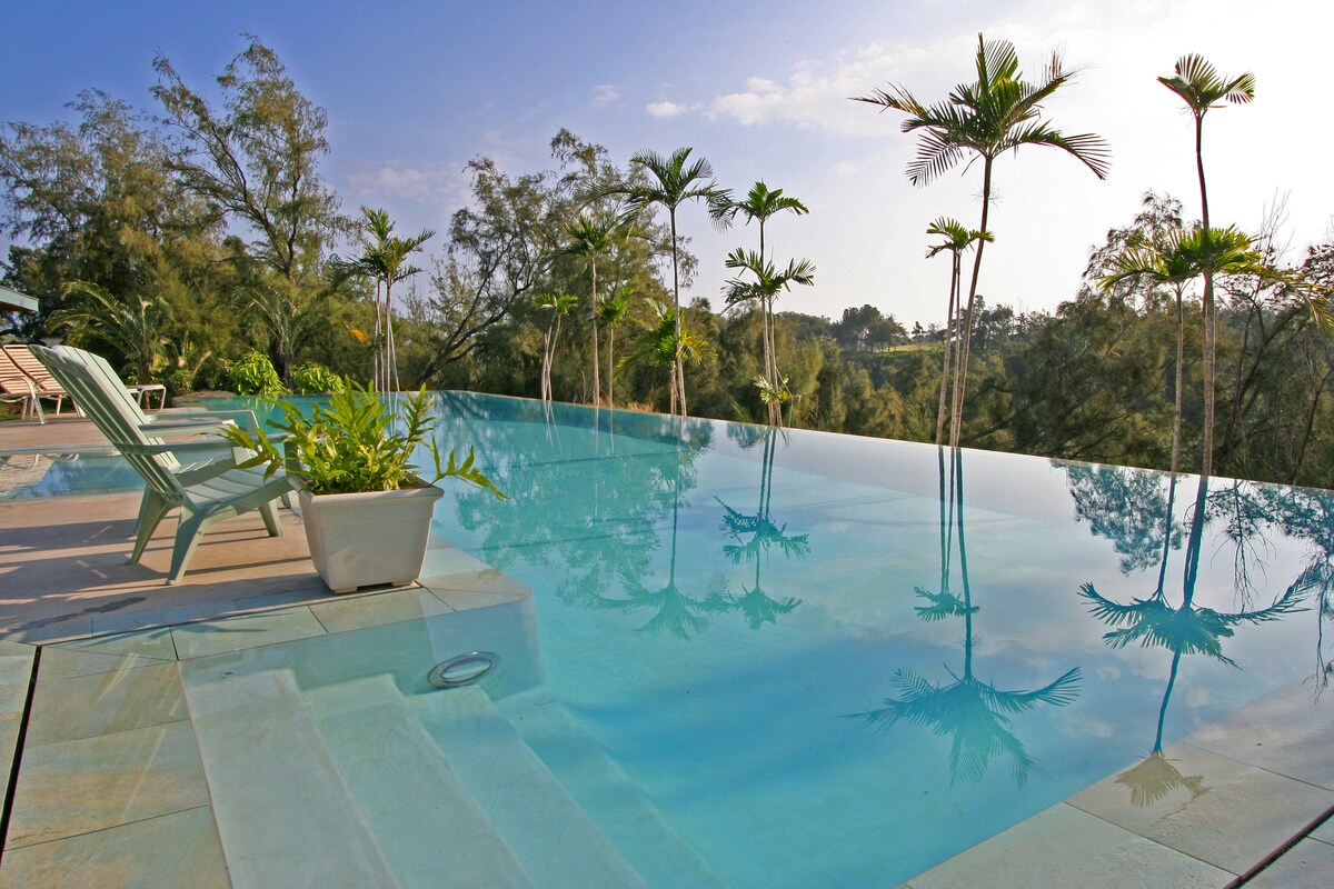 An infinity pool extends into the landscape, featuring a smooth transition from shallow steps to deeper waters. Surrounding palms reflect gently on the water's surface. Lounge chairs are positioned nearby, offering a serene spot for relaxation with views of the natural surroundings.