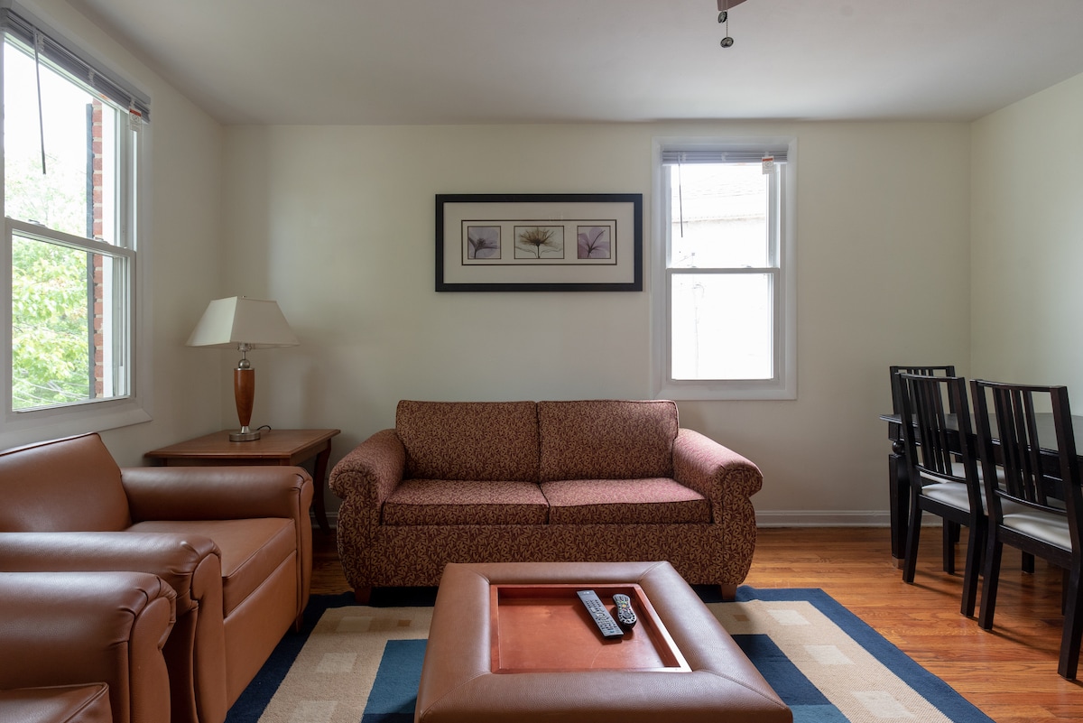 A welcoming living area includes a patterned brown sofa paired with two leather armchairs. Natural light filters through two windows with simple white blinds. A small wooden side table holds a lamp, and a rectangular coffee table occupies the center, enhancing the space's functionality.