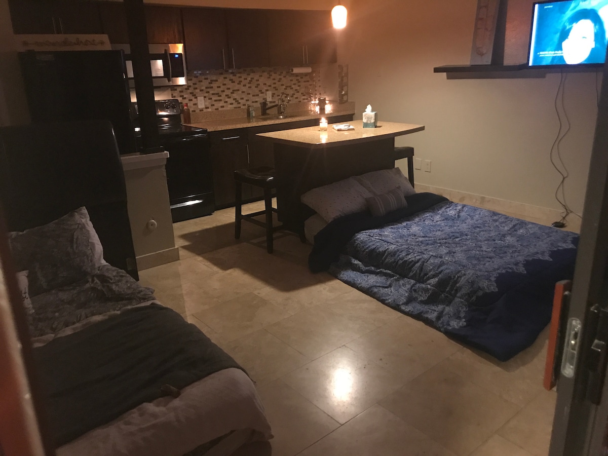 A vintage studio space is presented with two neatly arranged bedding setups on the tile floor. A contemporary kitchen area is visible in the background, featuring dark cabinetry and a mosaic backsplash. A small dining table is seen near the center, with a television mounted on the wall.