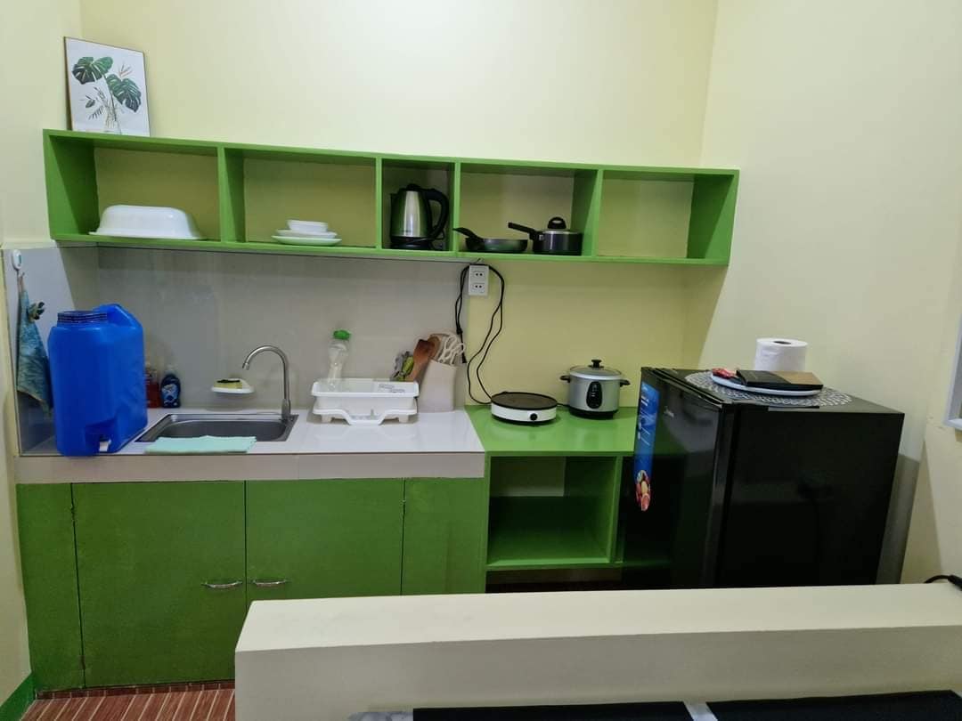 A compact kitchen area features green cabinetry with open shelves displaying a kettle and small kitchenware. A sink is positioned beside a white dish rack, while a rice cooker sits on a countertop next to a black refrigerator. A spacious dining table is partially visible in the foreground.