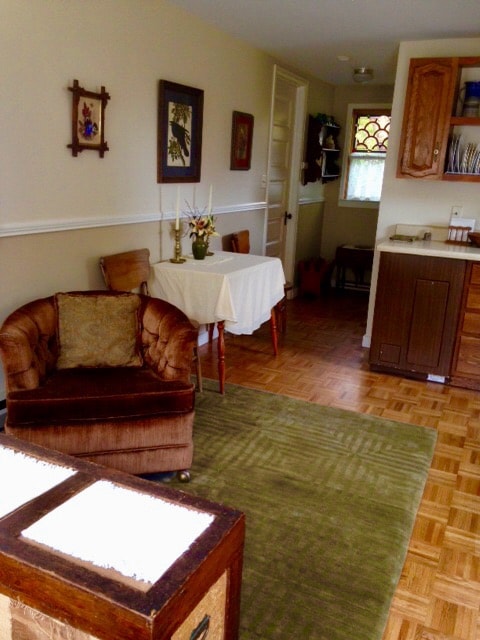 A living area presents a comfortable armchair beside a small table, adorned with a vase of flowers. Light wood cabinets are visible in the background, complemented by a corner view of the kitchen. The warm flooring features a soft green rug.