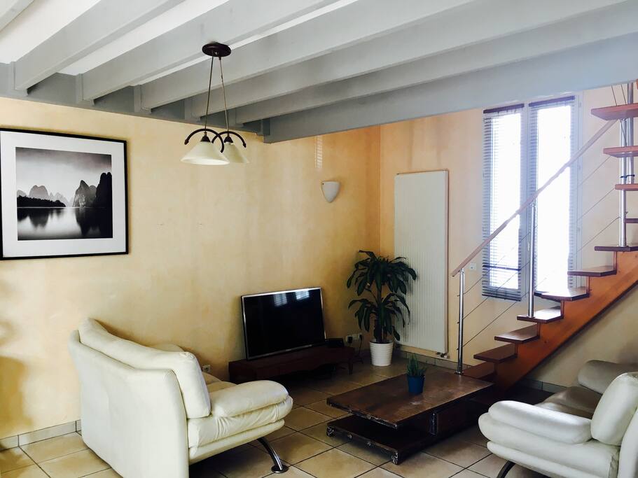 Partie salon avec accès à l'étage. Poutres apparentes, 4 mètres de plafonds de chaque côté. Traversant avec une fenêtre côté rue (sur photo) et deux grandes portes fenêtre donnant sur le patio privé de 80m2.