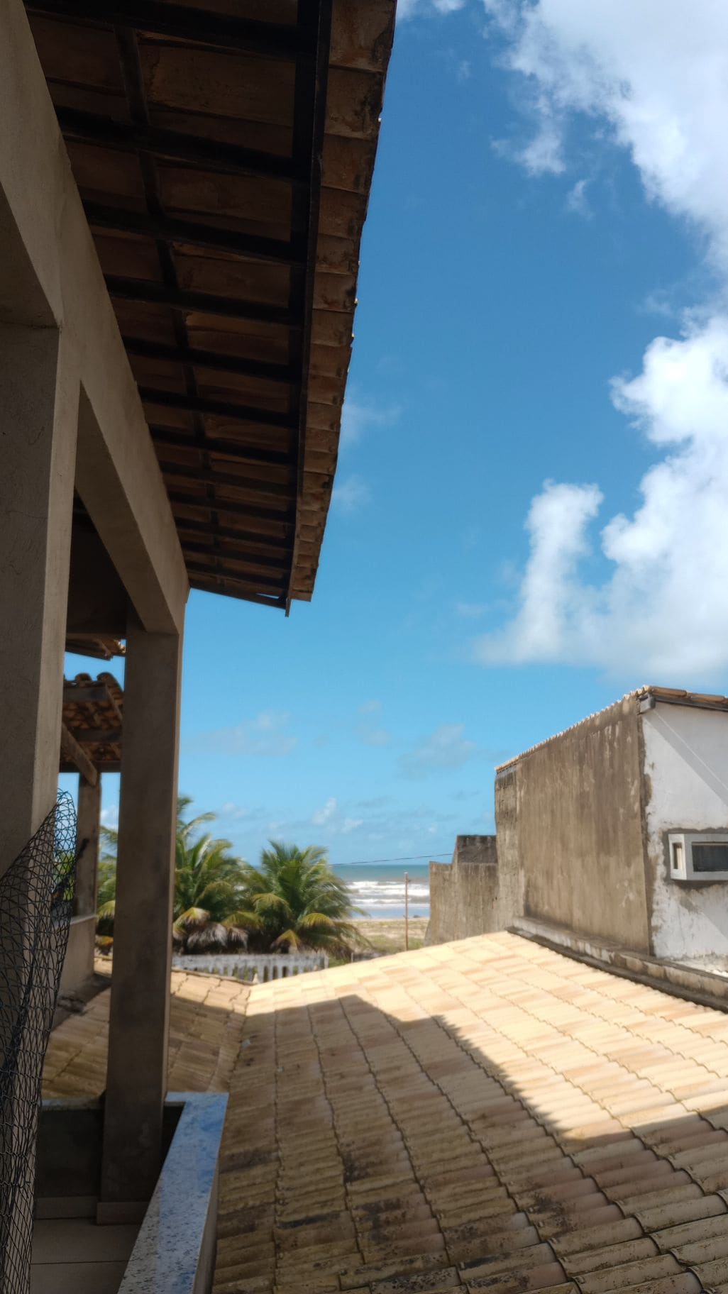 A view of the beach is visible from a rooftop terrace, featuring a tiled roof and clear blue skies. Palm trees can be seen in the foreground, while the ocean waves gently roll onto the shore in the background.